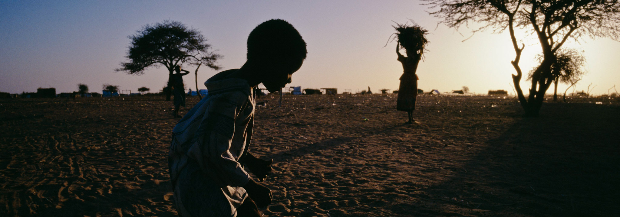 Silhouette of a child crosses in front of the camera with a silhouette of a woman carrying a bundle on her head in the background (Darfur, Sudan)
